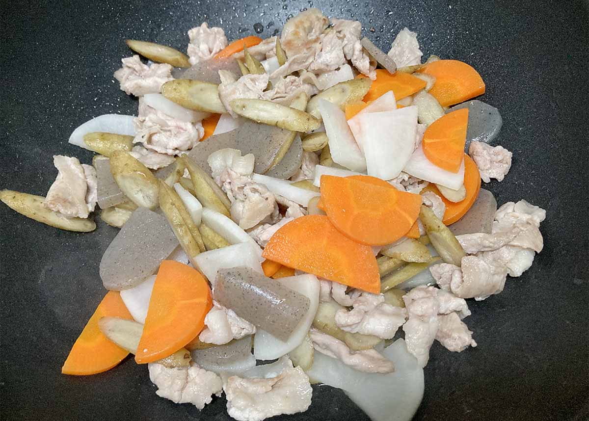 Stir-fry of pork with carrot slices, onion pieces, and pale root vegetables in a dark pan.