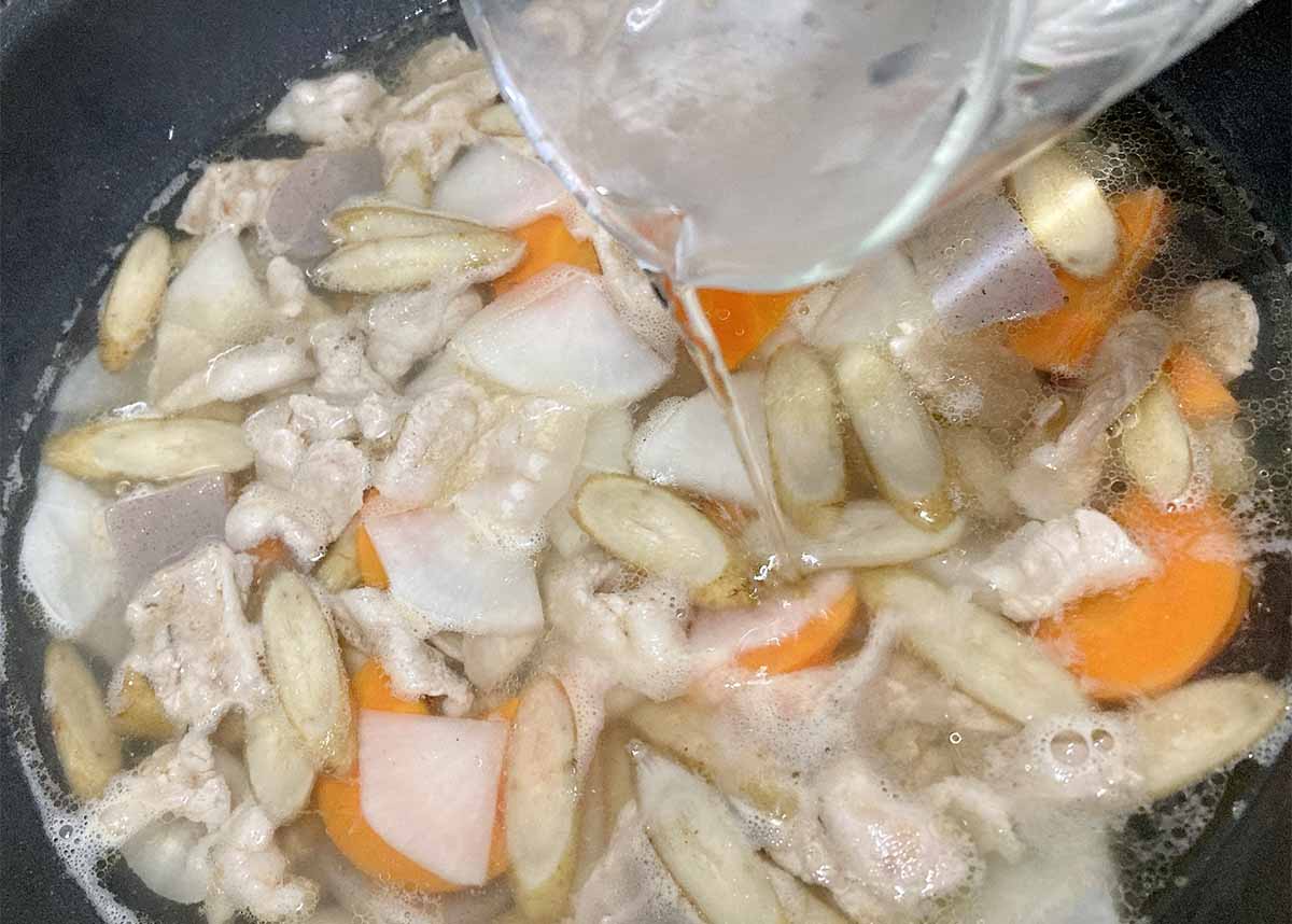 Pot of chicken stew with carrots and potatoes simmering as water is poured in.